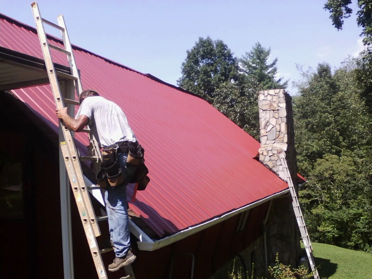 Roofer on a ladder beside a red metal roof and stone chimney — Soffit & Fascia Repair by Roofing Brothers in Pico Rivera